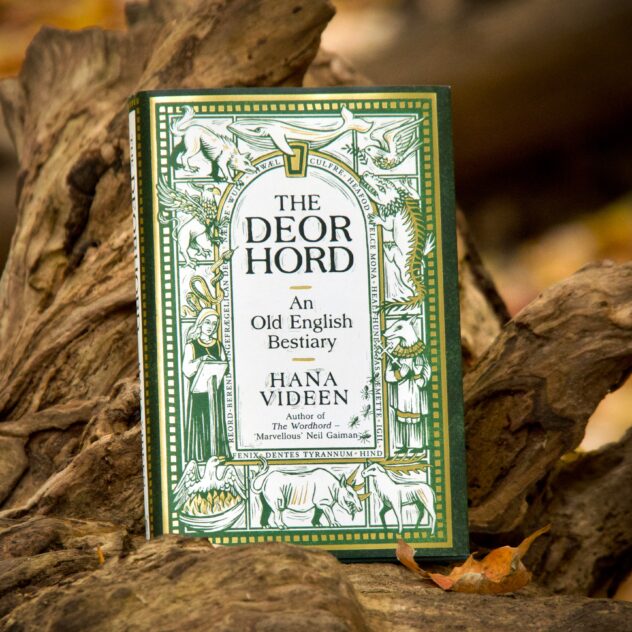 A book, “The Deorhord: An Old English Bestiary”, by Hana Videen, resting on a log with an orange leaf beside it. The cover background is white with a gold and green border with decorative gold squares. The title and author’s name are written inside an arched window shape, with a border of words around it: culfre, heafod swelce mona, healf-hundingas, æmette, igil, hind, dentes tyrannum, fenix, reord-berend, ungefrægelican deor, nædre, wulf and hwæl. The area surrounding the Old English word border has wood-cut style illustrations in green and gold, each in its own compartment but overlapping slightly: a wolf, a whale, a dove, a spiky lizard with big teeth, a dog-headed person holding a cross, a deer, a weird-looking rhino-like creature, a phoenix in a burning nest, a nun writing, a snake and a griffin. By the author’s name it says “Author of The Wordhord”, which Neil Gaiman described as “Marvellous”, and some ants crawl alongside it.